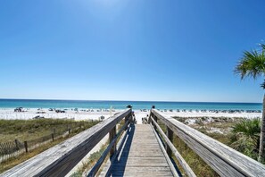 Ein Holzsteg führt zu einem Strand mit blauem Himmel und ruhigem Wasser.
