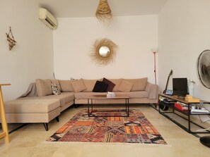 Een woonkamer met een beige bank, een bijzettafeltje, een kamerkleed met dessin, een wandairconditioner, een hanglamp en een staande ventilator.