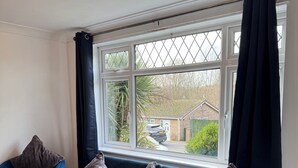A blue sofa with four cushions in a room with a large window and a view of a residential area. - Stylish 3-Bed with Smart TVs, Parking & Workspace (Kettering)