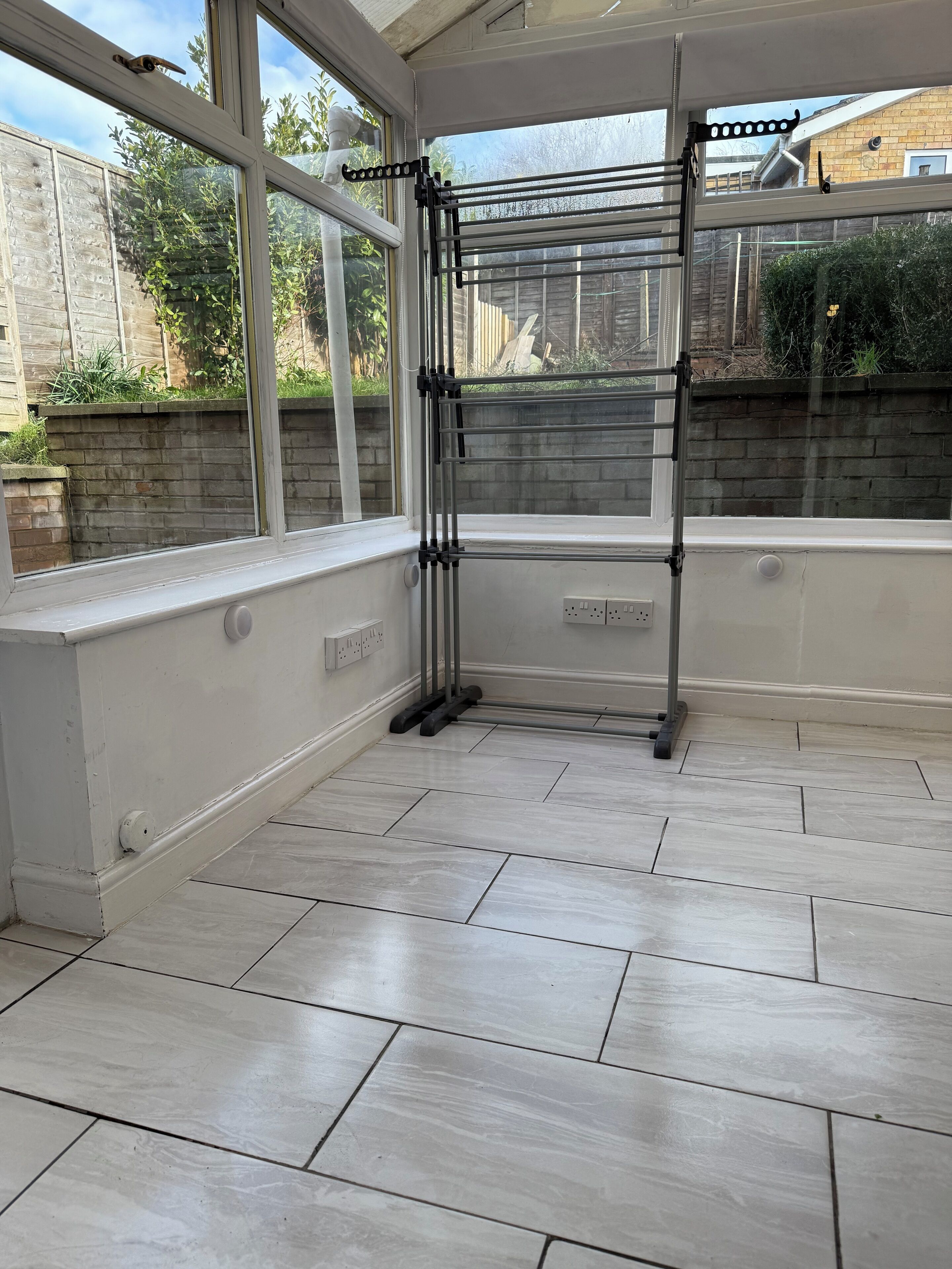 A conservatory with a built-in drying rack, tiled floor, and large windows offering a view of the garden and neighboring buildings.