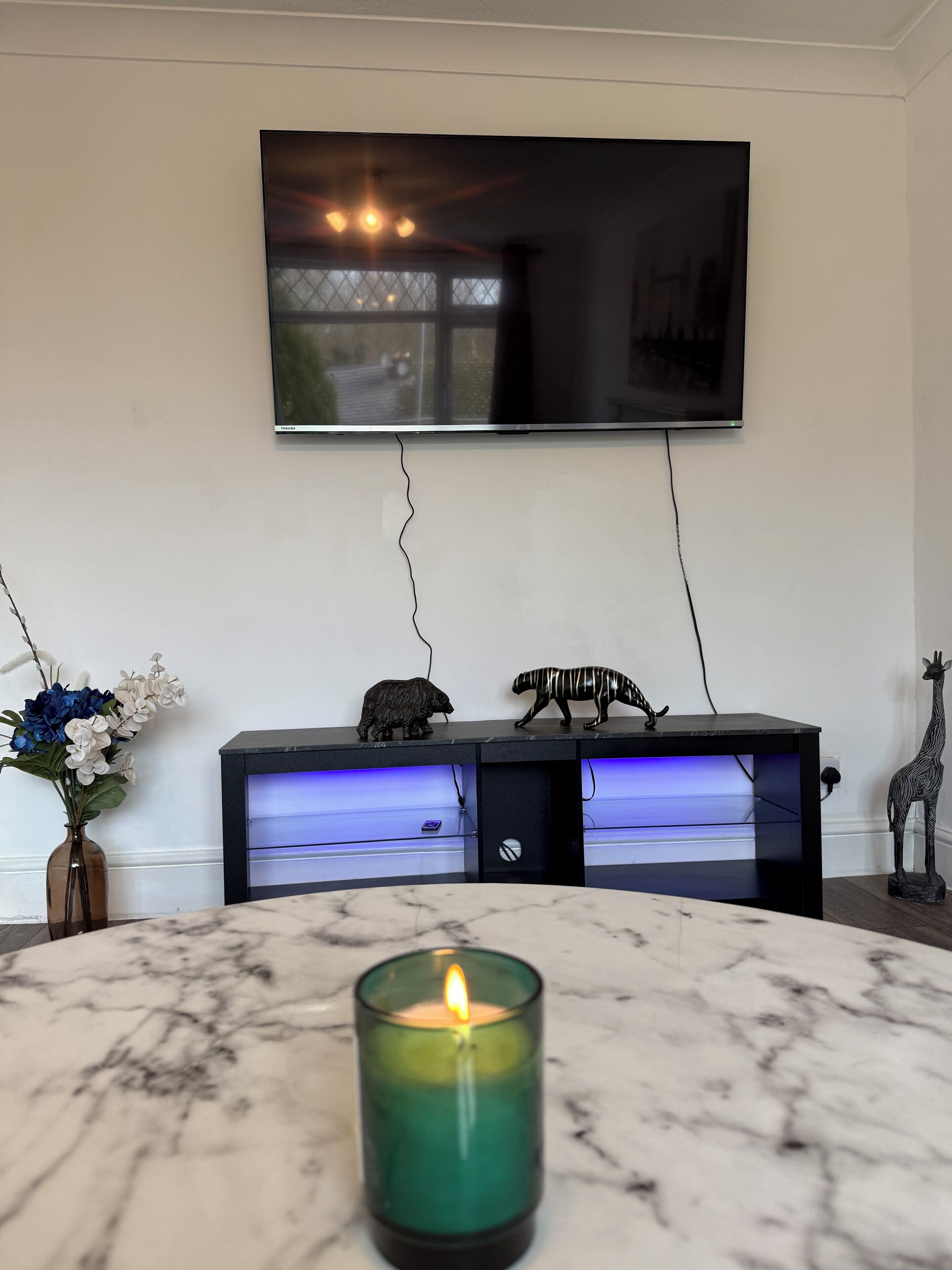 Een woonkamer met een flatscreen televisie aan de wand, een marmeren bijzettafeltje, een brandende kaars en decoratieve objecten op een zwarte console tafel.