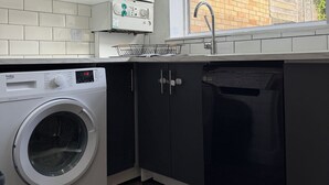 A modern kitchen with a built-in washer and dryer, a window with a view of a brick wall, and a sink with a faucet. - Stylish 3-Bed with Smart TVs, Parking & Workspace (Kettering)
