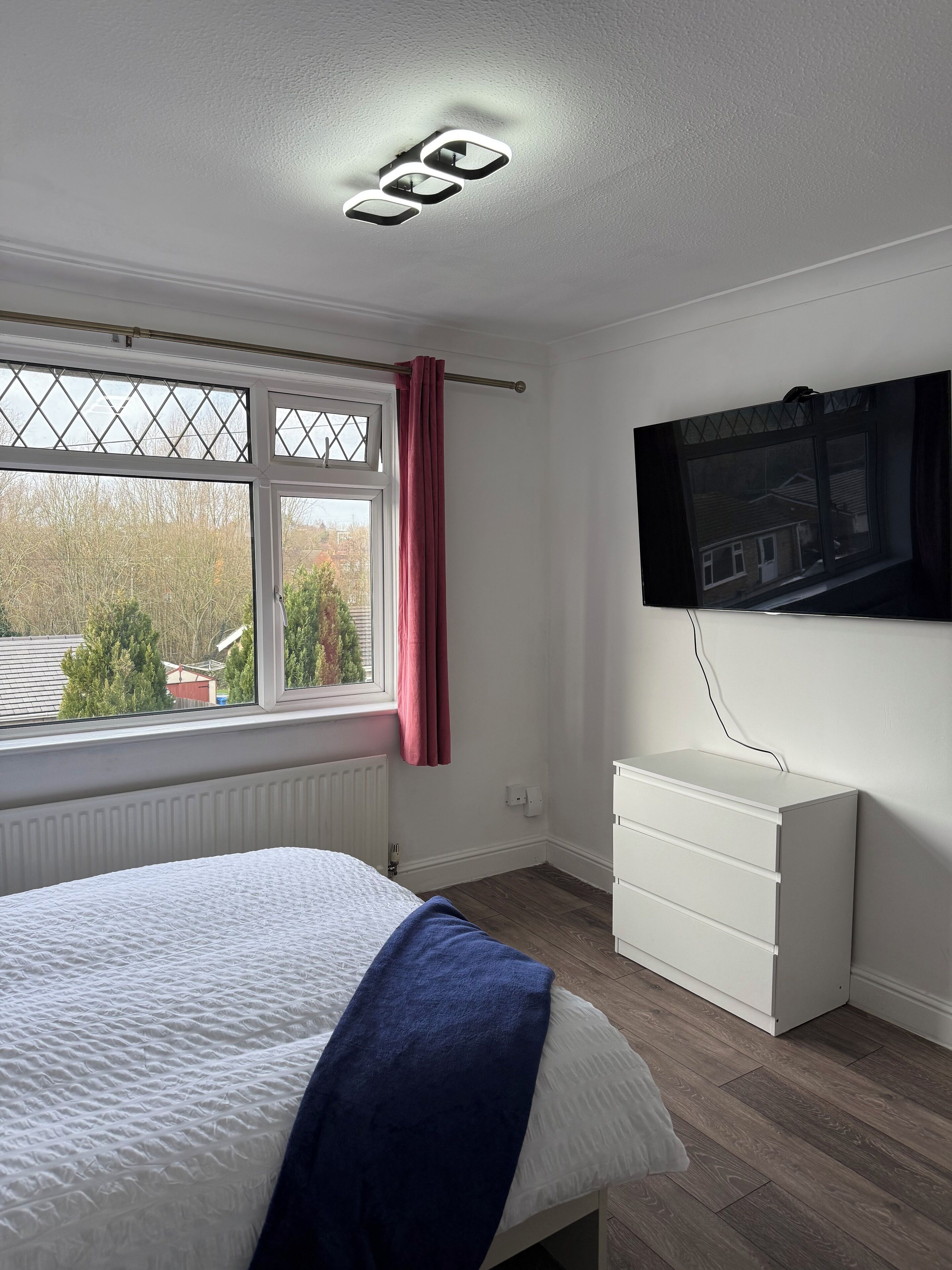 Een slaapkamer met een bed, een raam met uitzicht, een radiator, een witte dressoir en een aan de muur bevestigde televisie.