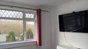 Een slaapkamer met een bed, een raam met uitzicht, een radiator, een witte dressoir en een aan de muur bevestigde televisie.