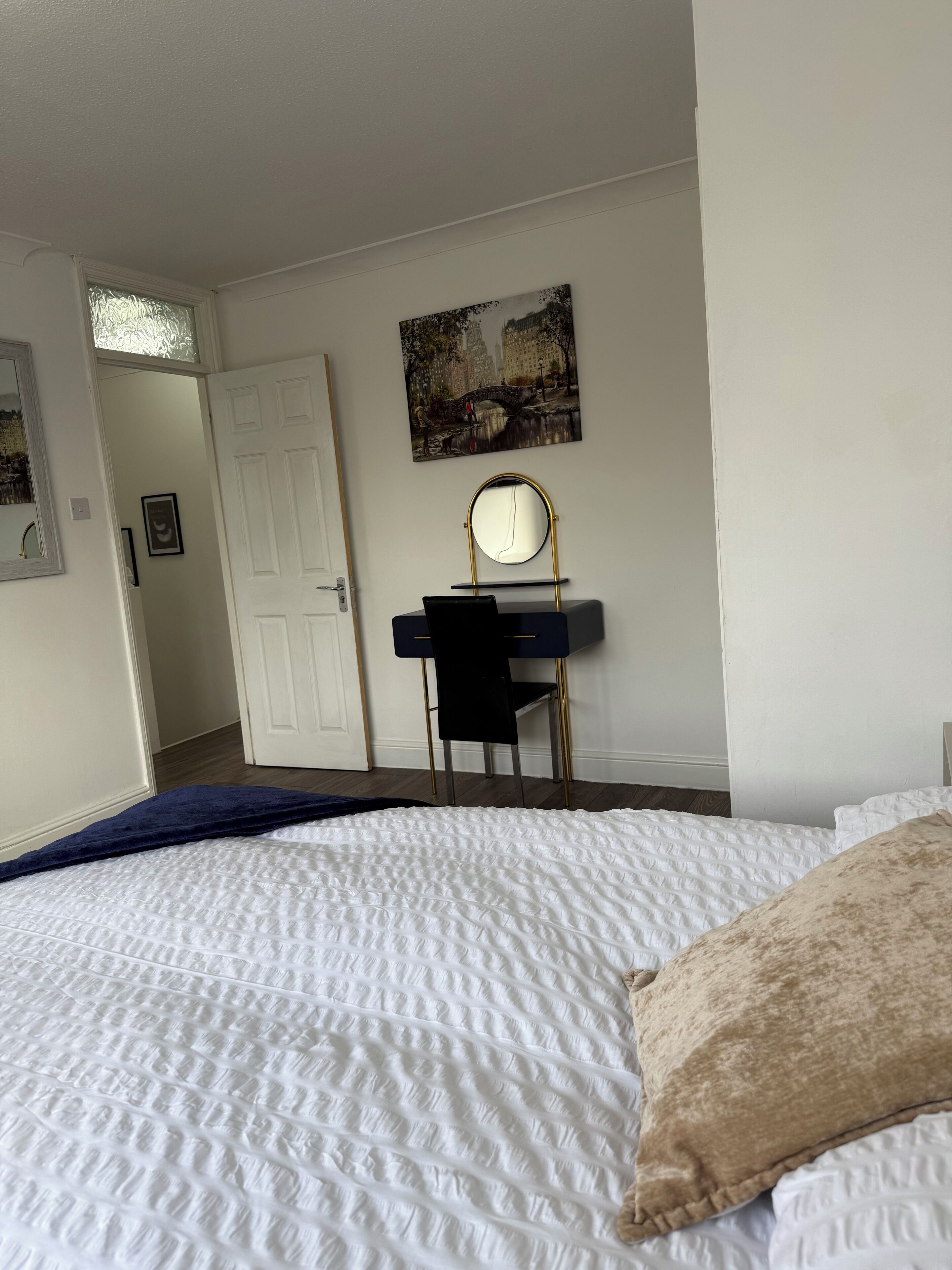 A bedroom with a bed, a desk with a mirror, a door, and a wall-mounted picture.