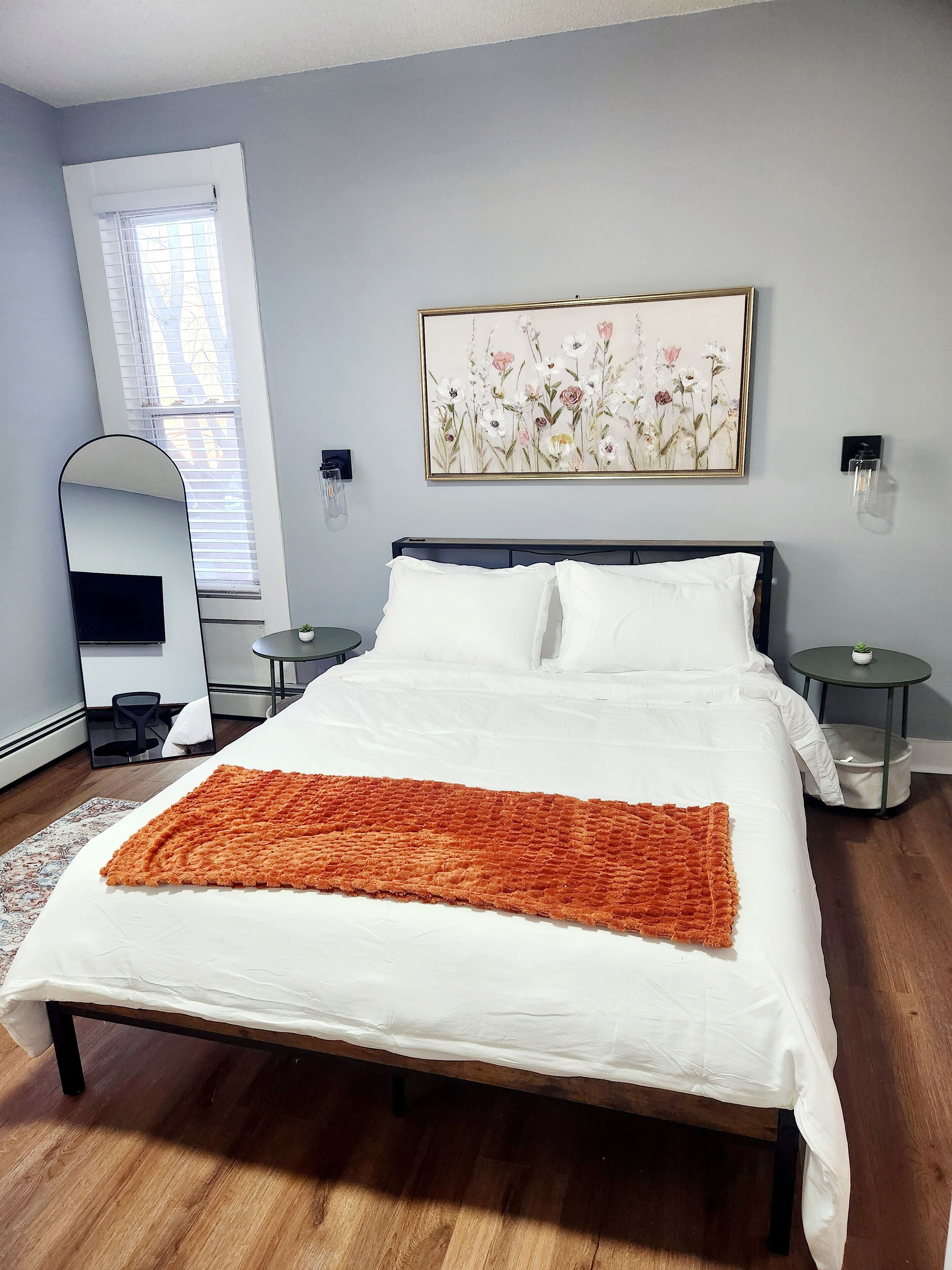 A bedroom with a large bed, a bedside table, a small round table, a mirror, a window with blinds, and a floral artwork on the wall.