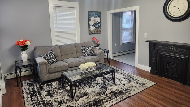 Un salon avec un canapé gris, des coussins à motifs noir et blanc, une table basse en verre, un tapis noir et blanc, une cheminée et une horloge murale.