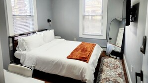 Une chambre à coucher comprenant un lit, deux fenêtres avec stores, une table de chevet, une lampe et un téléviseur fixé au mur.