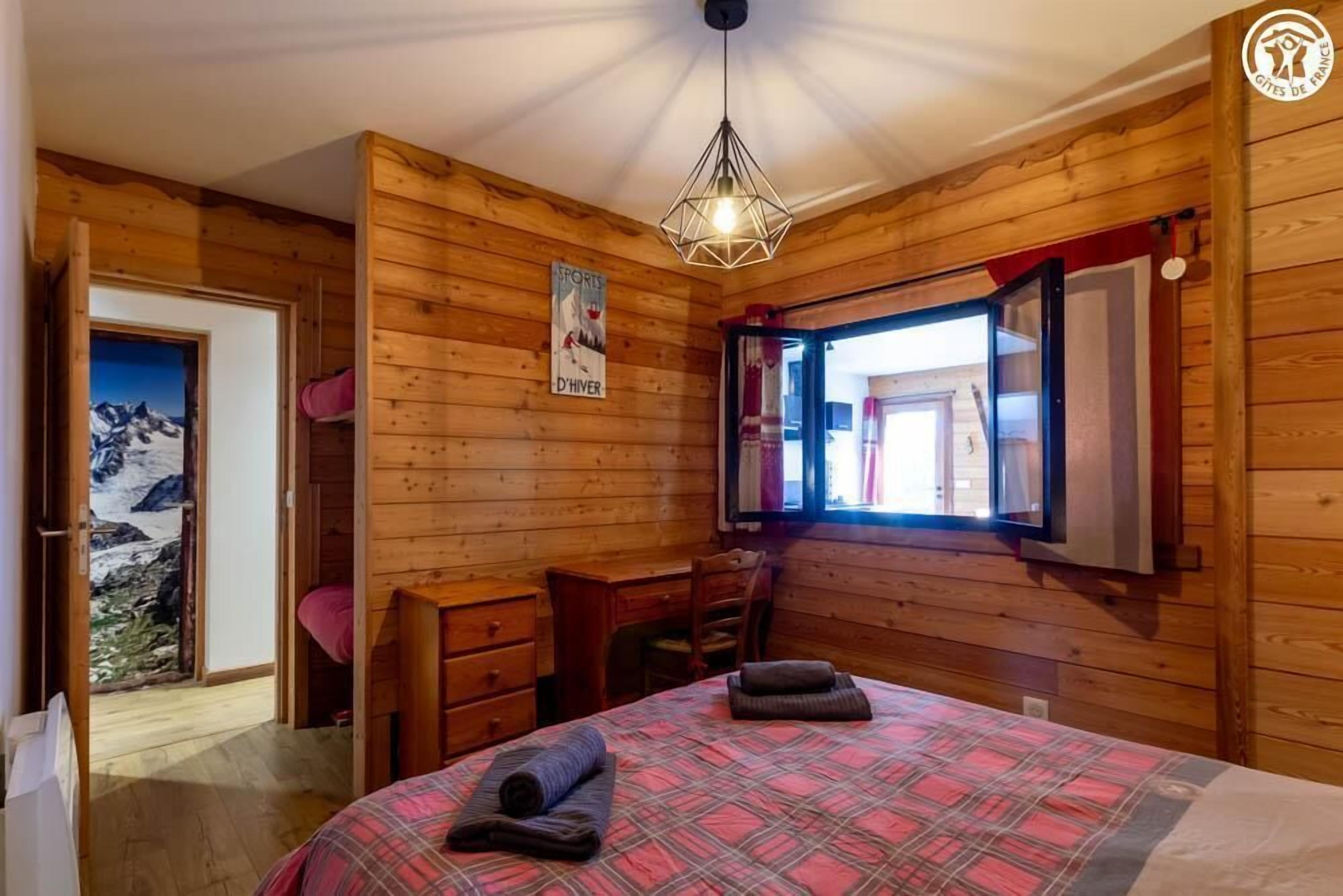 A wooden-paneled room with a bed, a desk, and a chair. There is a window with a view of the outdoors.