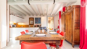 A dining area with a wooden table set for six, surrounded by red chairs. A kitchen is visible in the background with modern appliances. - GITE DES FONTANETTES (LA COMPOTE)