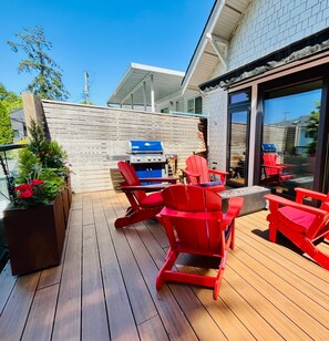 Een houten terras met rode Adirondack-stoelen, een barbecue en een groot raam.