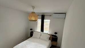 Ett sovrum med en säng, sängbord, en hängande lampa, ett fönster med persienner och en luftkonditioneringsanläggning.