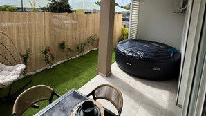 En uteplats med ett bord dukat för två personer, ett bubbelbad och utsikt över en trädgård med staket och grönska.