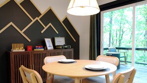 A dining area with a round table, four chairs, a cabinet with items, and a view of trees through a window.
