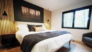 A modern bedroom with a large bed, a nightstand, a side table, a window with a view of trees, and two wall-mounted picture frames.