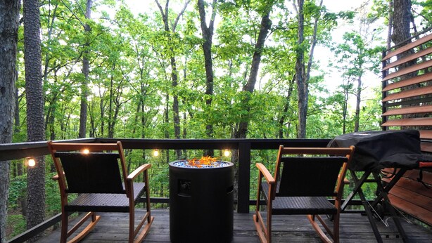A deck with two rocking chairs, a round table with a fire pit, and a view of a forest.