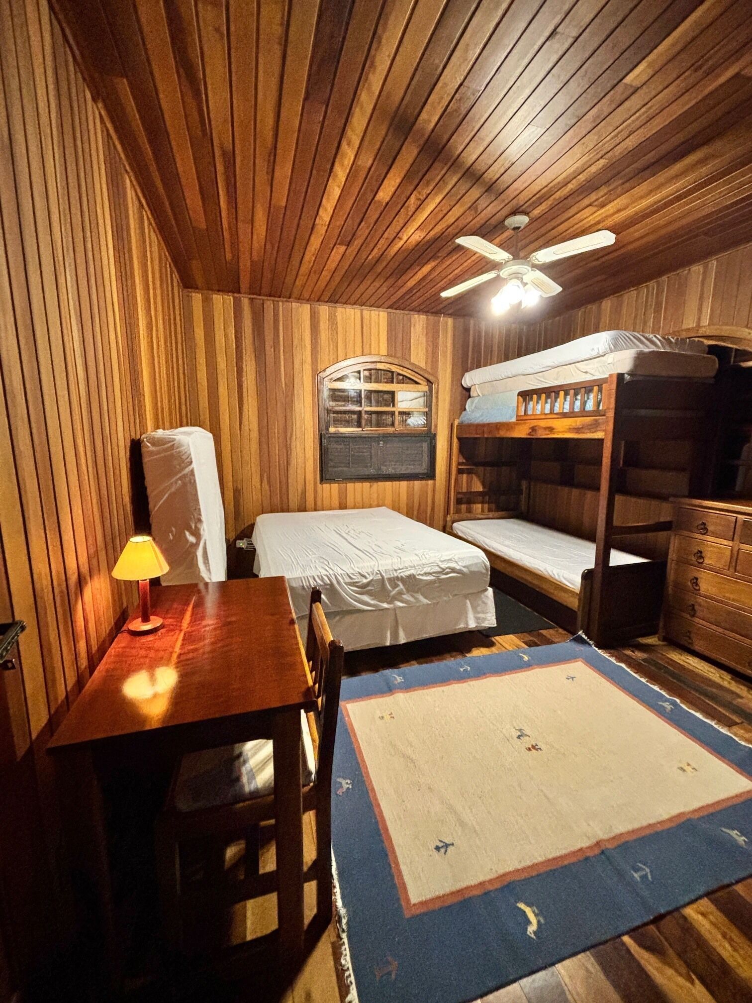 A cozy room with wooden paneling, a bunk bed, a desk with a lamp, and a window with a view.