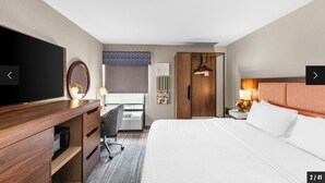 A hotel room with a bed, a desk with a chair, a television, a wardrobe, and a window with blinds.