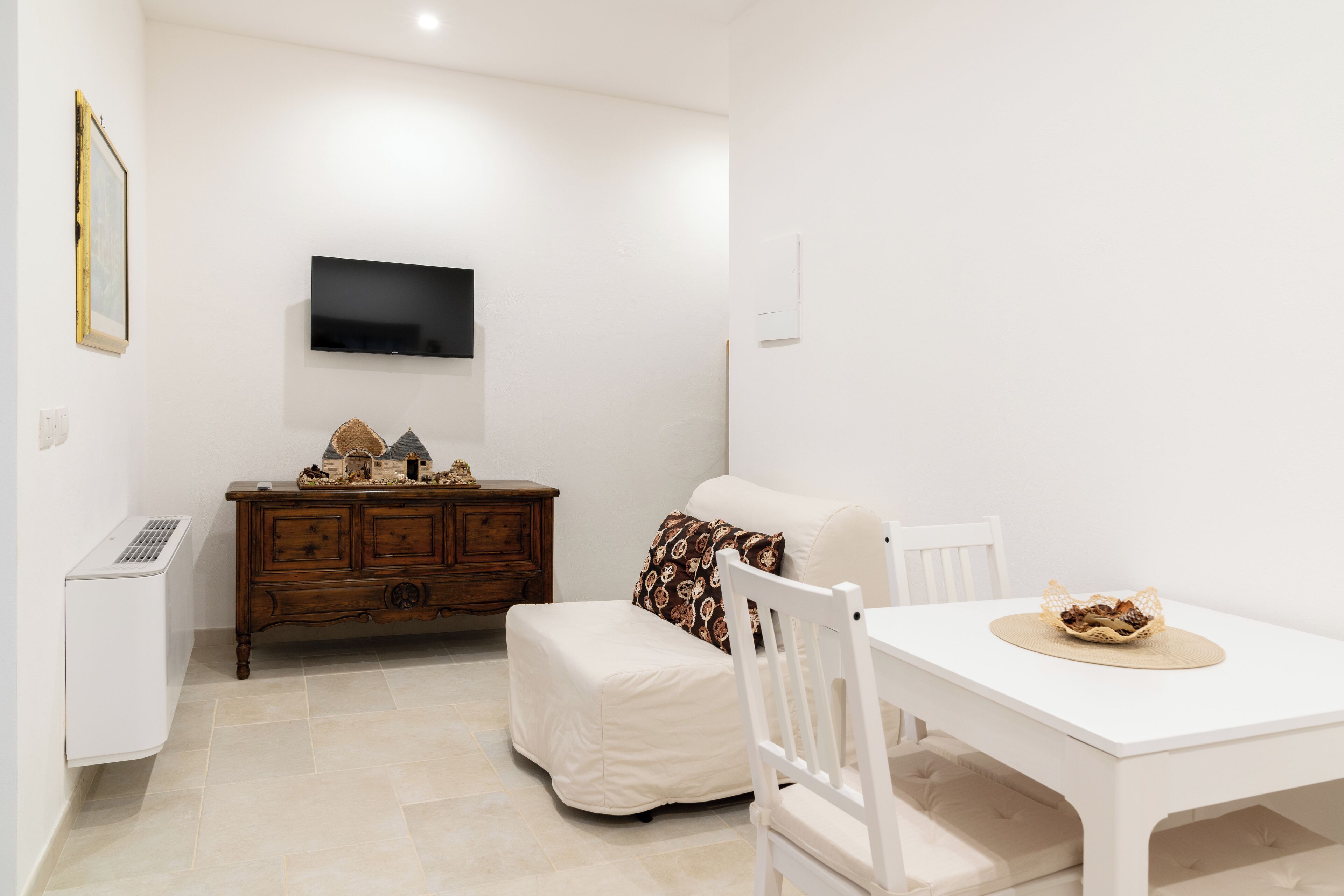 A small, clean room with a white table, a white sofa, a wooden dresser, and a flat-screen TV mounted on the wall.