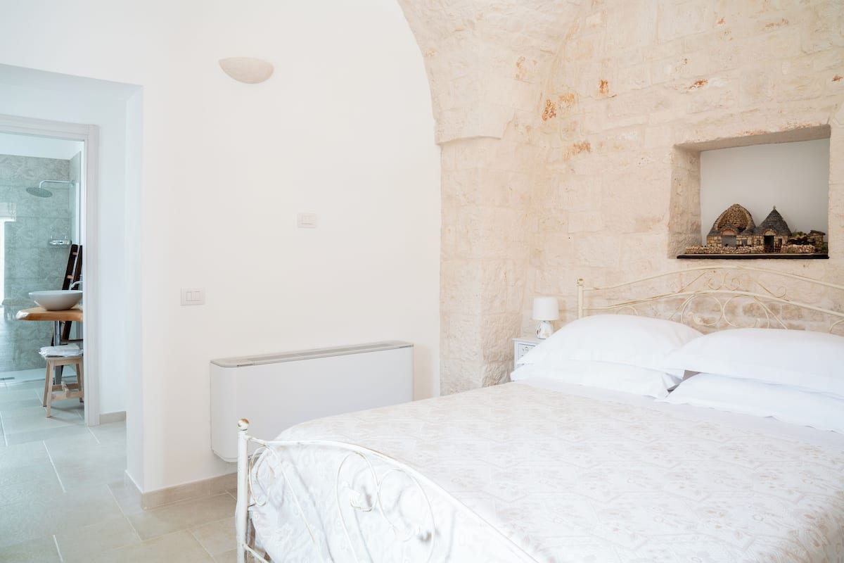 Une chambre à coucher avec un lit, une table de chevet, une étagère ornée d’objets décoratifs et un mur en pierre.
