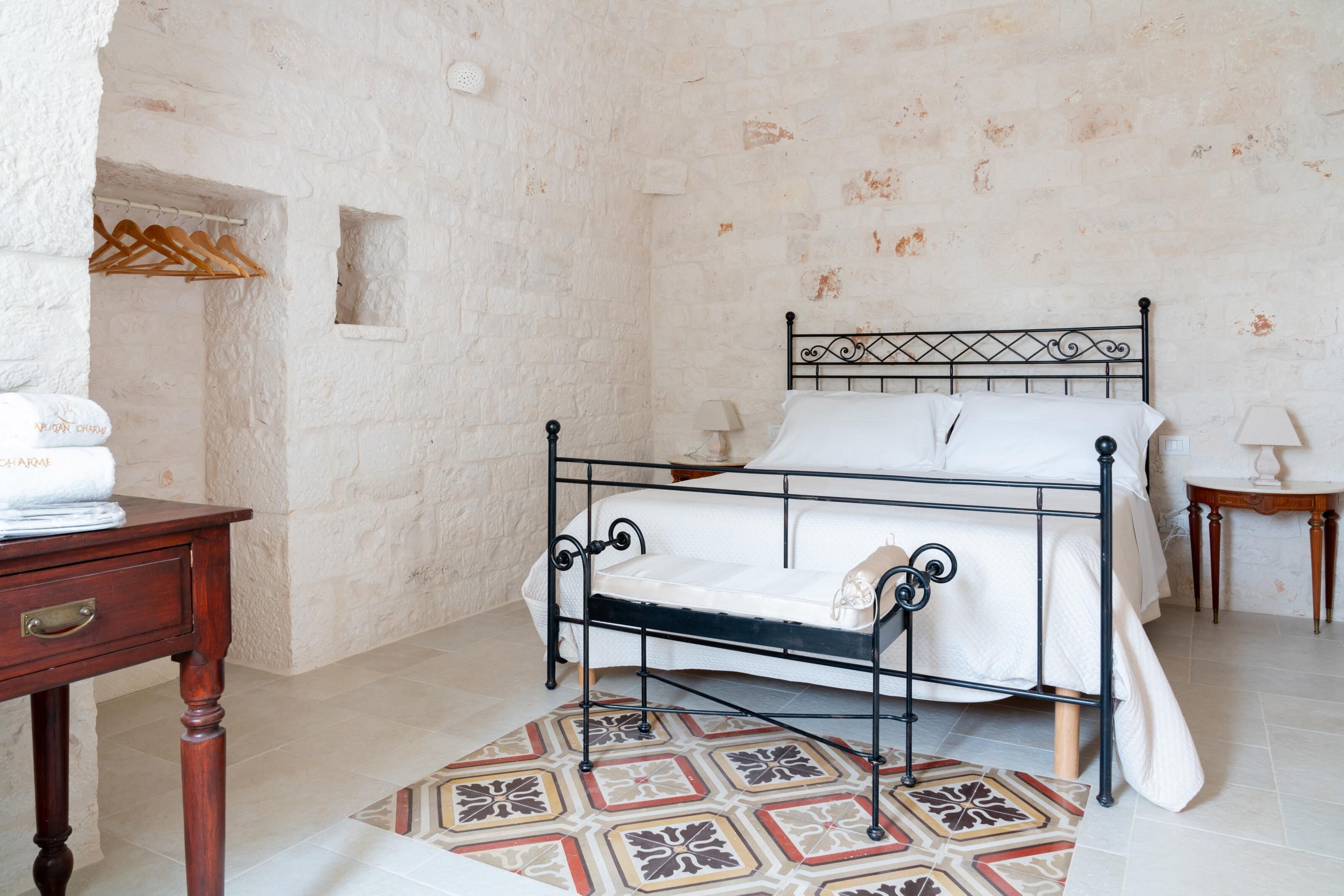 Een slaapkamer met een metalen bedframe, witte beddengoed, een kamerbreed tapijt met een patroon, een houten nachtkastje en een kleine tafel met een lamp.