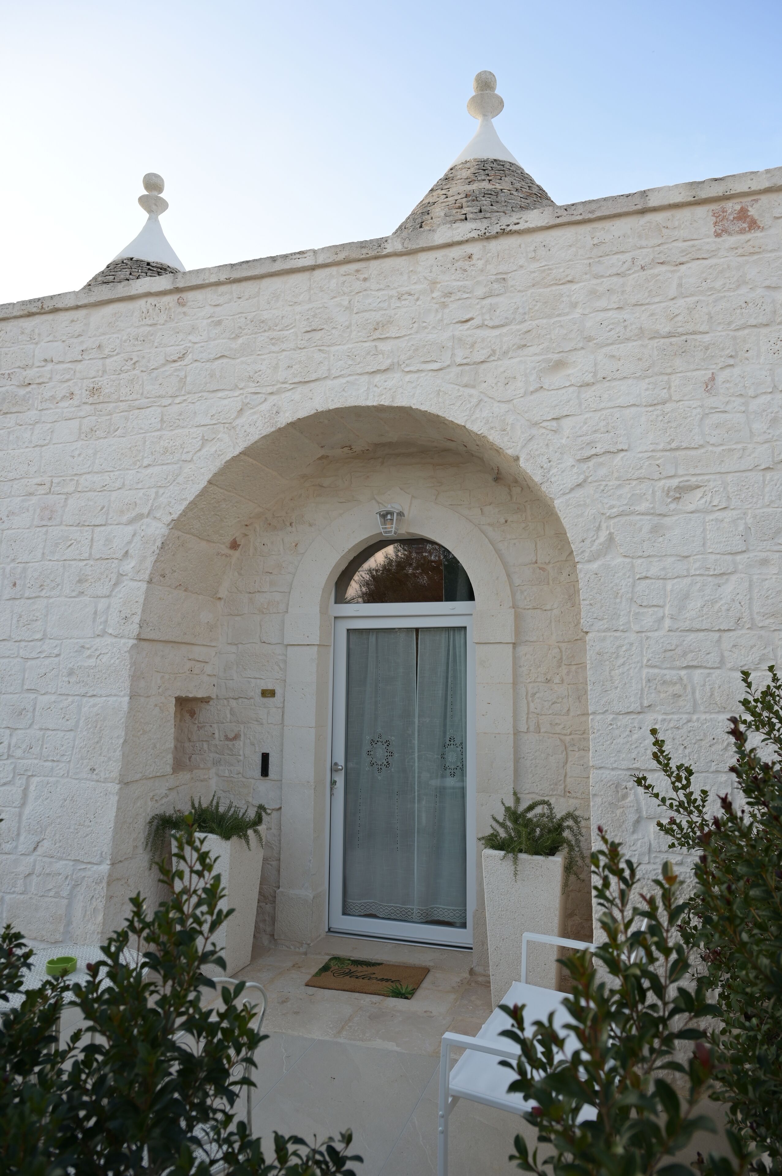 Un edificio de piedra blanca con una entrada arqueada, una puerta con panel de vidrio y un pequeño balcón con una silla y plantas en macetas.