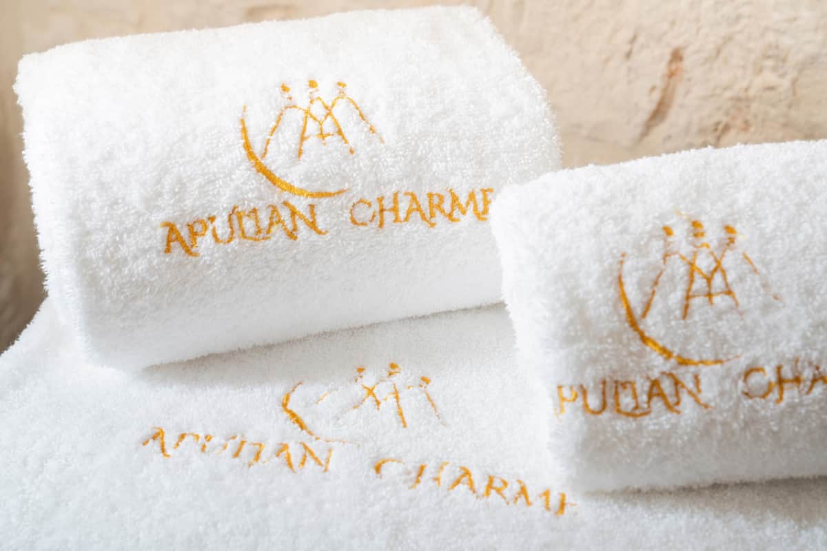 Trois serviettes blanches ornées de motifs brodés et d’un logo, disposées sur une surface texturée.