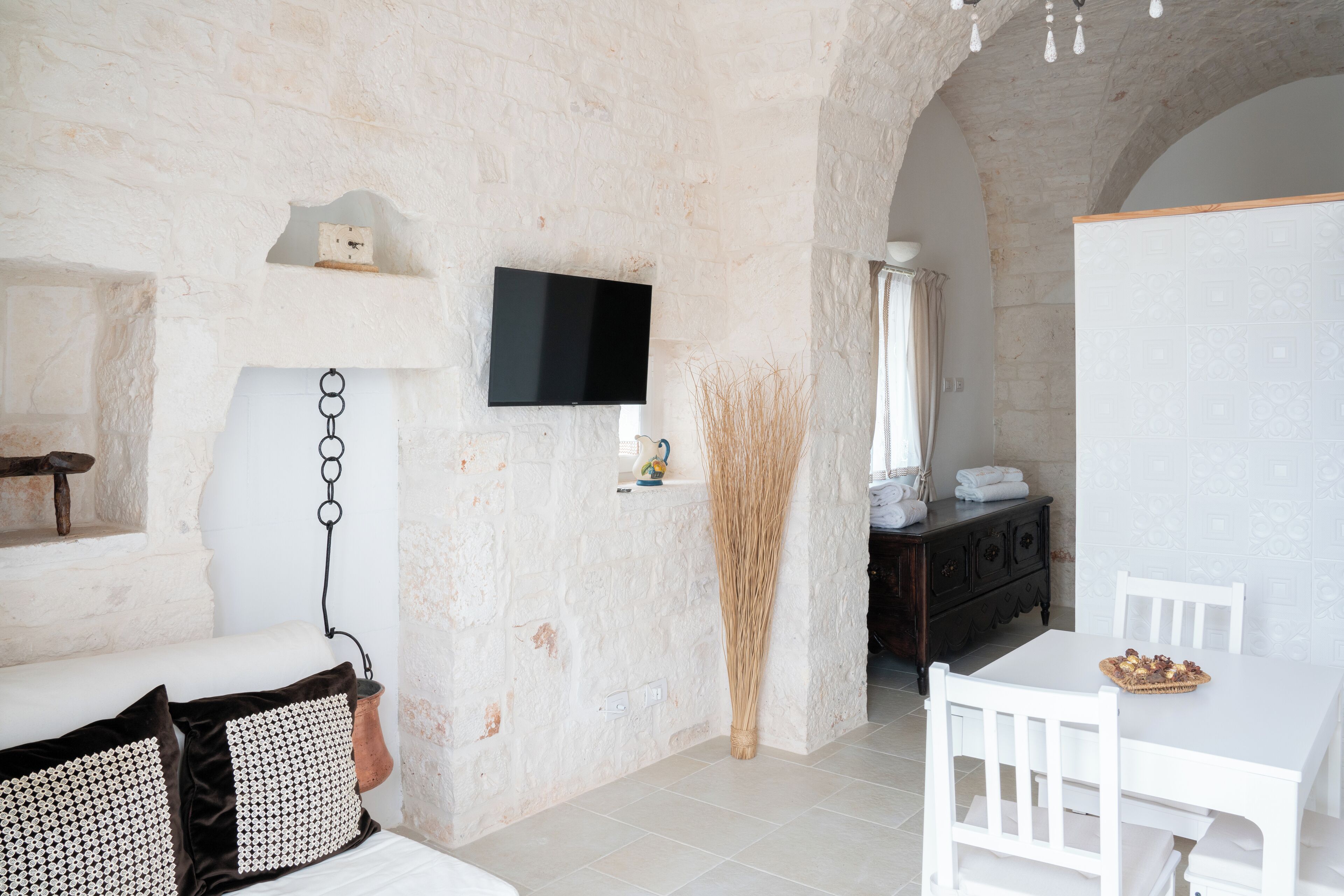 Une pièce avec un lit blanc, une table blanche avec des chaises, un téléviseur fixé au mur et un mur en pierre rustique.