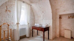 Una habitación con pared de piedra rústica, una silla de madera, una pequeña mesa de madera con toallas y una ventana con cortinas blancas.