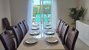Un espace repas avec une longue table dressée pour le repas, des poutres en bois au plafond et une vue sur un espace extérieur verdoyant, visible à travers une porte vitrée.