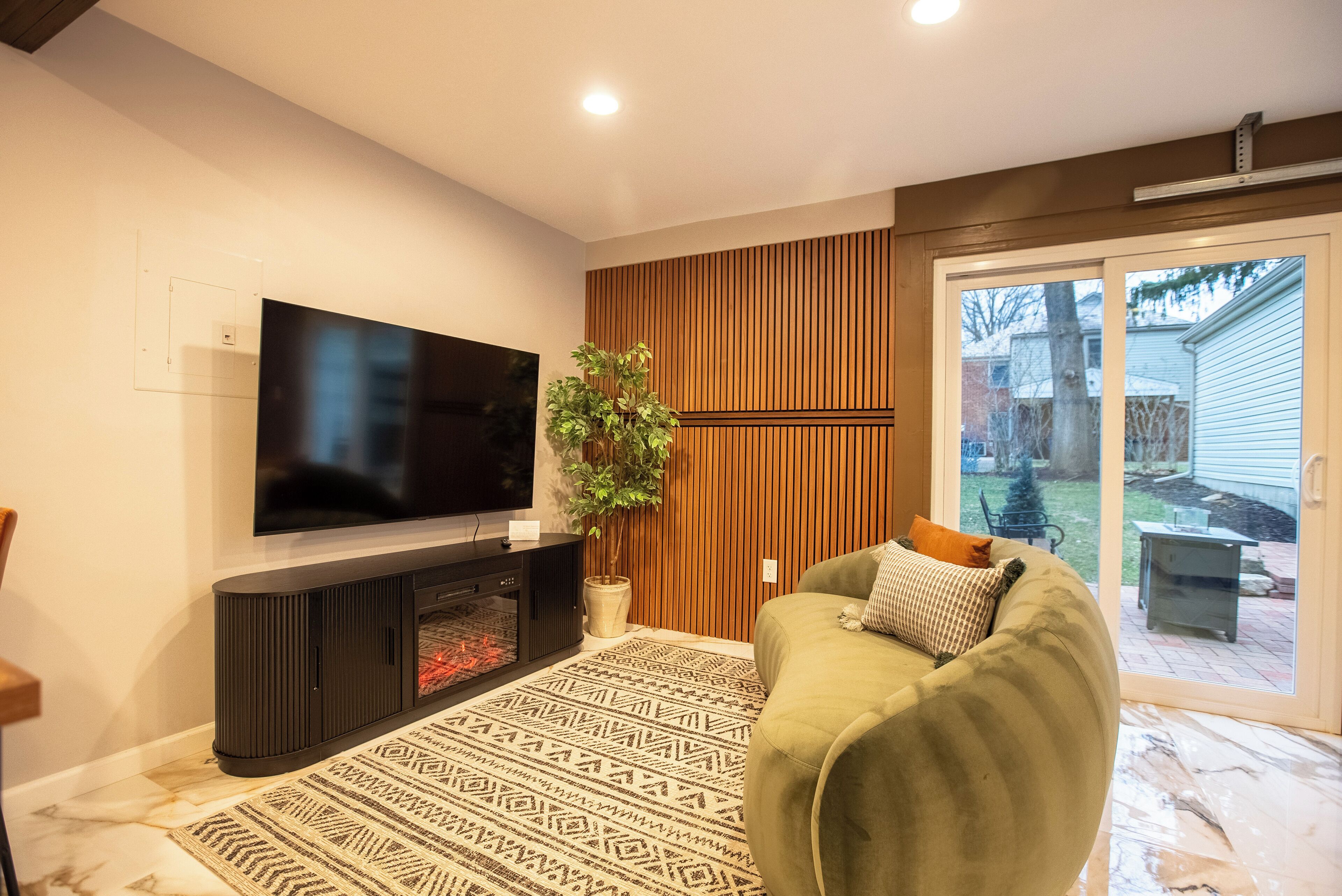 Ein Wohnzimmer mit Flachbildfernseher, einer dunklen Unterhaltungseinheit, einem gemusterten Teppich, einem grünen Sofa und einer Topfpflanze.