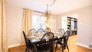Un coin repas avec une table en bois dressée pour six personnes, entourée de chaises noires, d’un lustre et d’une fenêtre agrémentée de rideaux.