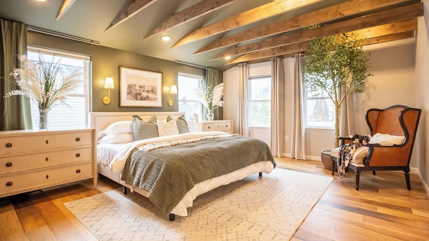 Une chambre à coucher avec un grand lit, des poutres en bois, une table de chevet, une chaise et une fenêtre avec des rideaux.