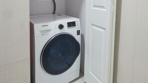 A compact laundry room with a front-loading washing machine, a small electrical outlet, and a door leading to another room. - Luxurious 2BR/2BTH APT in New Kingston (Kingston)