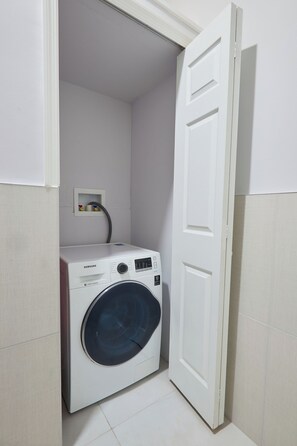 A compact laundry room with a front-loading washing machine, a small electrical outlet, and a door leading to another room. - Luxurious 2BR/2BTH APT in New Kingston (Kingston)