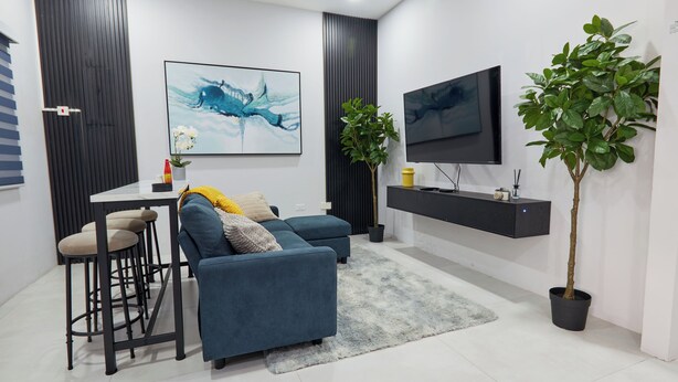 A modern living room with a blue sofa, a dining table with stools, a flat-screen TV, a potted plant, and artwork on the walls. - Luxurious 2BR/2BTH APT in New Kingston (Kingston)