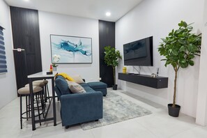 Un salon moderne avec un canapé bleu, une table à manger avec des tabourets, un téléviseur à écran plat, une plante en pot et des œuvres d’art accrochées aux murs.