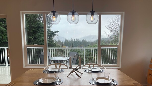 Ein Essbereich mit einem Holztisch, gedeckt für vier Personen, große Fenster mit Blick auf Bäume und Berge sowie Pendelleuchten darüber.