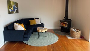 Ein Wohnzimmer mit einem dunkelblauen Sofa, einem runden Holztisch, einem gemusterten Teppich, einem schwarzen Kaminofen und einer hängenden Leuchte.
