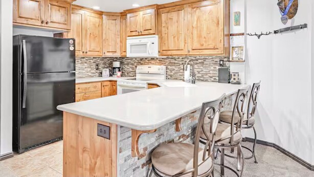 A kitchen with wooden cabinets, a black refrigerator, a white microwave, and a stone backsplash. - Stay at Giant Steps, Ski in / Ski out, Sleeps 4, Full Kitchen (Brian Head)