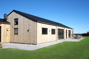 A modern wooden building with a sloped roof, large windows, and a patio area with outdoor furniture. - Nice holiday home 100 meters from the water (Rudkøbing)