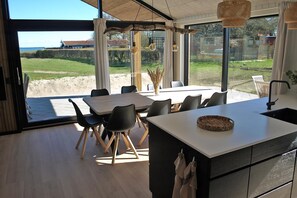 Une cuisine moderne dotée d’un grand îlot central, d’un coin repas avec des chaises et de grandes fenêtres offrant une vue sur l’extérieur.