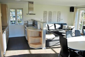 A modern kitchen with a built-in stove, a dining area with a black leather sofa and white table, and a large window with a view of trees. - Holiday home just 400 meters from the water (Rudkøbing)