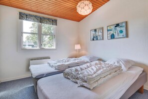 A bedroom with two beds, a window with a tree-patterned valance, a standing lamp, and two wall-mounted artworks. - Holiday home only 50 meters to the beach (Rudkøbing)