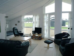 Een woonkamer met een televisie, lederen fauteuils, een houten tafel en grote ramen.