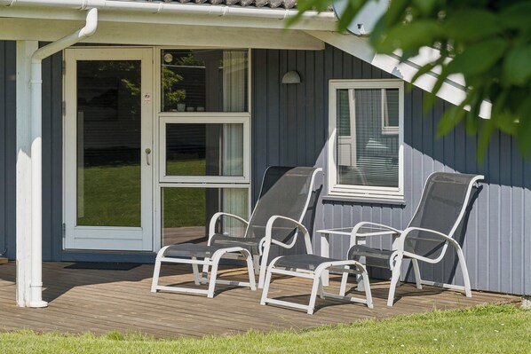 A covered deck with four white metal chairs and a table, a sliding glass door, and a window with blinds.