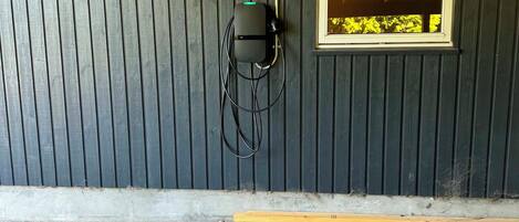 Een gevel met een donker houten wand, een raam met een geel kozijn, en een zwarte elektriciteitskast met kabels.