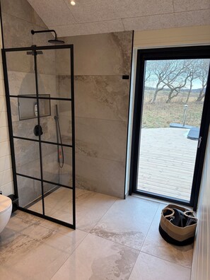A modern bathroom with a glass shower enclosure, a toilet, and a view of a wooden deck outside. - Lækkert sommerhus med udsigt over Limfjorden (Roslev)