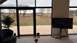 Una habitació amb grans finestres, una cadira blau fosc, una taula petita amb un objecte decoratiu i un televisor sobre un suport.