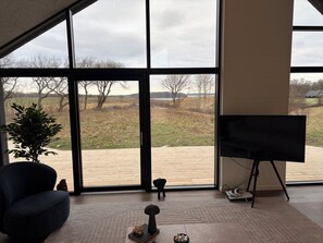 Ett rum med stora fönster, en mörkblå fåtölj, ett litet bord med ett dekorativt föremål och en tv-apparat på ett stativ.